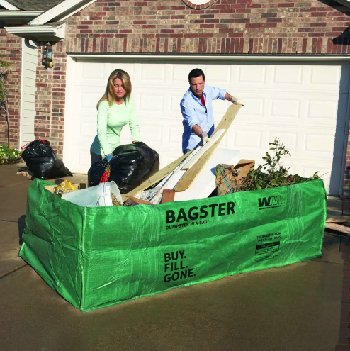 A husband and wife loading their junk into a large green bag that lays in their driveway as they donate.